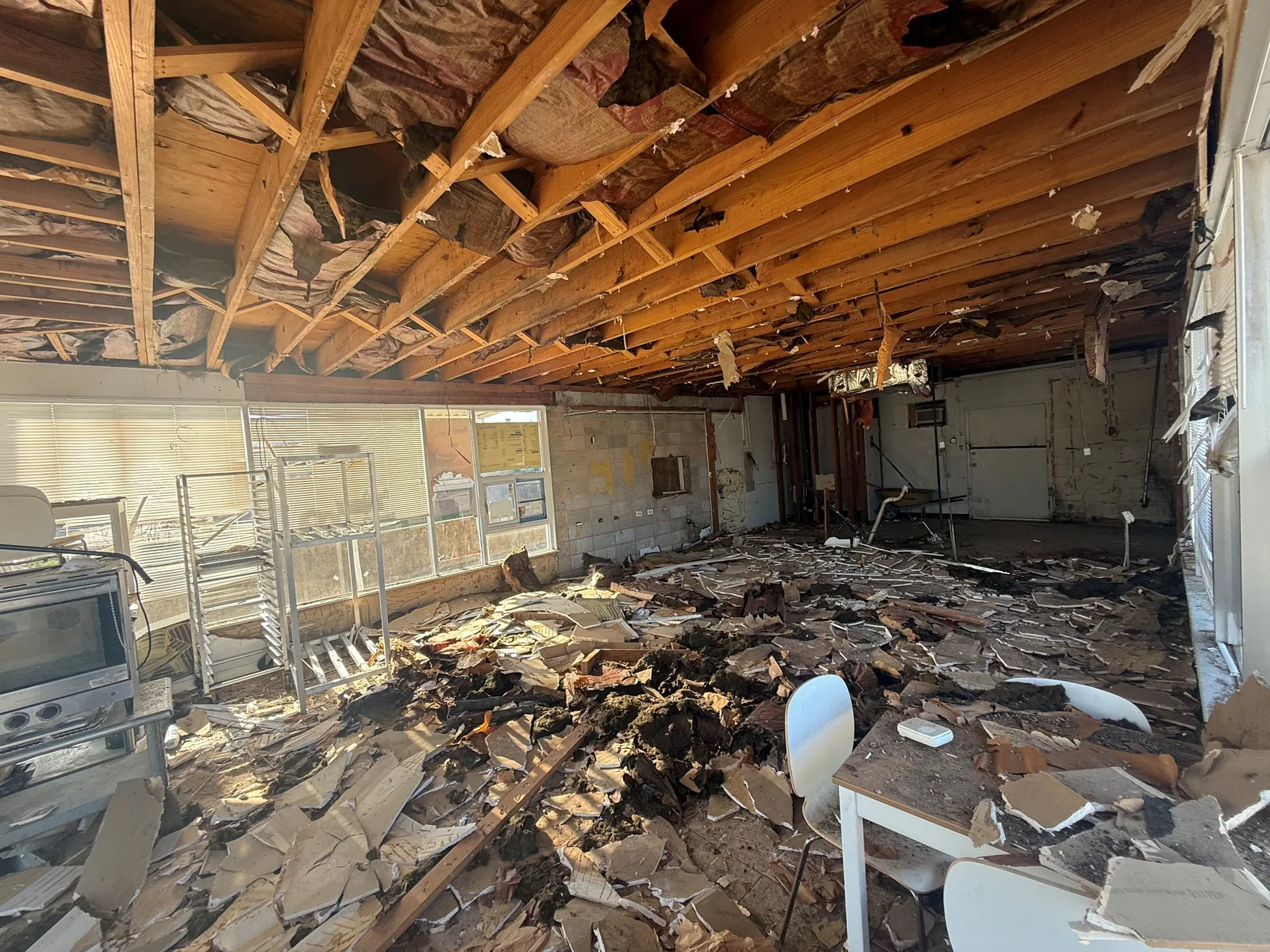 Demolition in full swing: the ceiling torn open and debris covering the floor, with the old bakery racks and oven still in place.